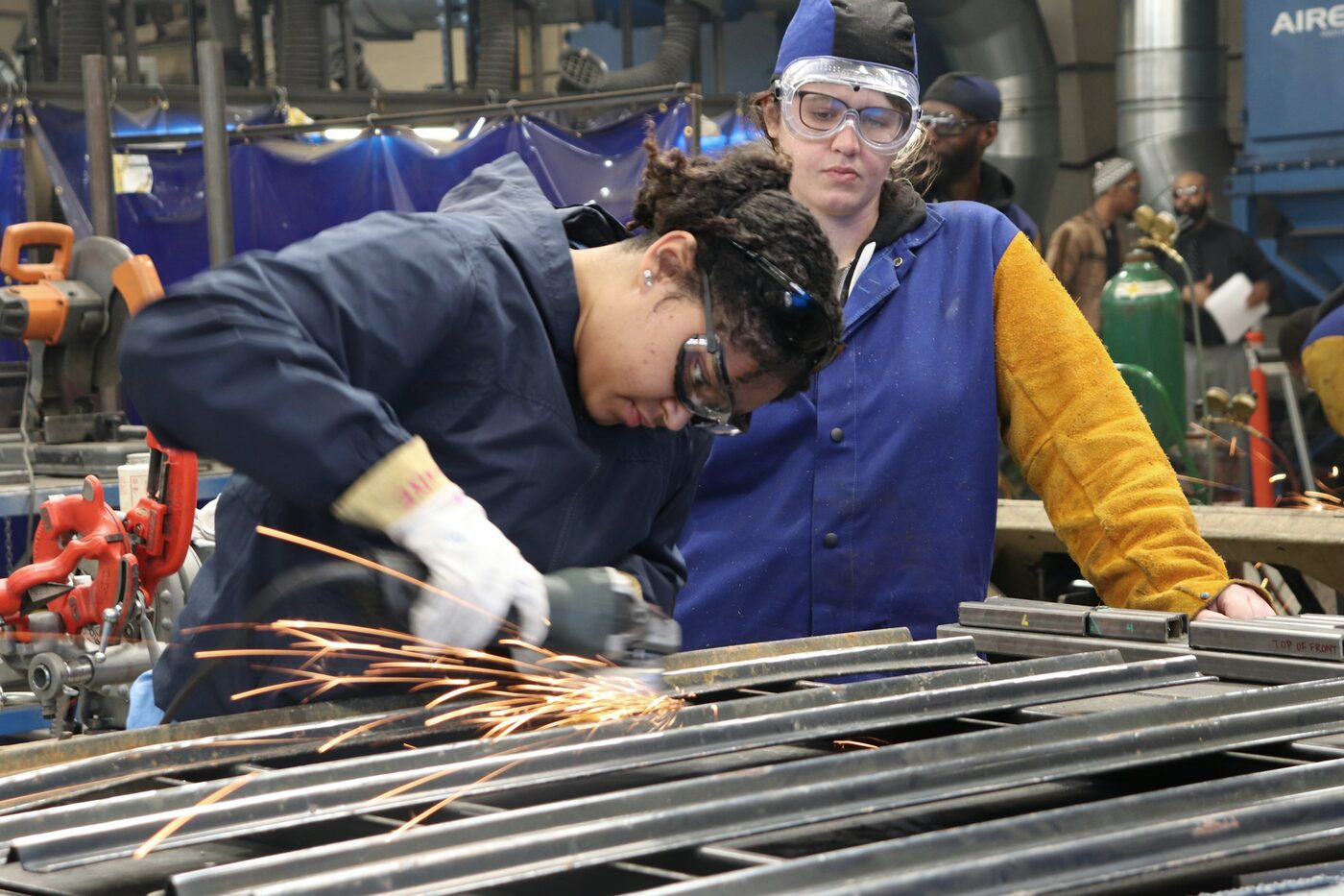 Two trainees in blue coveralls and face shields grinding steel in a training workshop