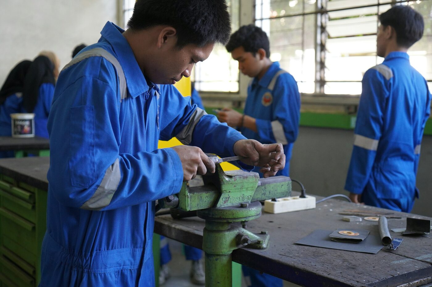 Trainees in blue overalls filing metal held in a bench vice